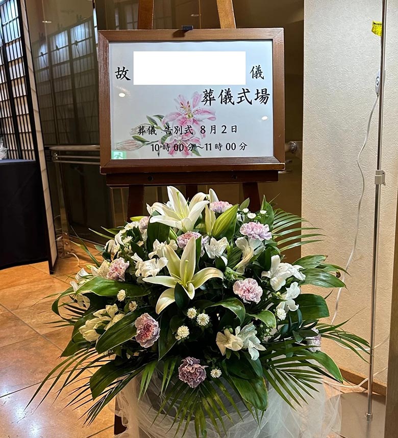 葬儀案内の看板と、その下には百合やカーネーションを使用した大きな花束が置かれています